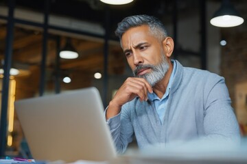 Middle-aged Hispanic man focuses intently on laptop computer, deep in thought while working in a modern office space during the day