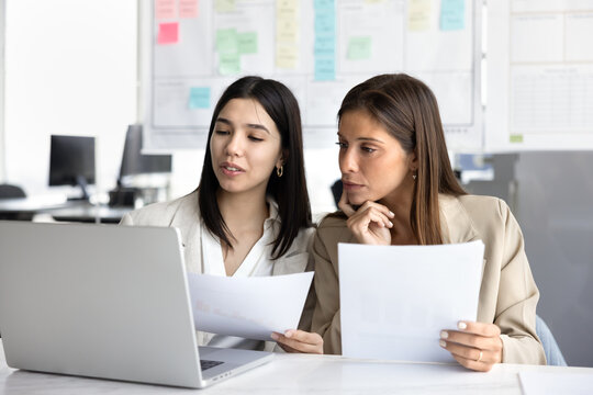 Two young diverse female coworkers sharing laptop