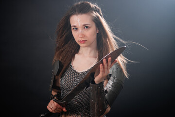 Naklejka premium Portrait of a young woman, fantasy warrior in armor with a sword in her hands isolated on a dark background