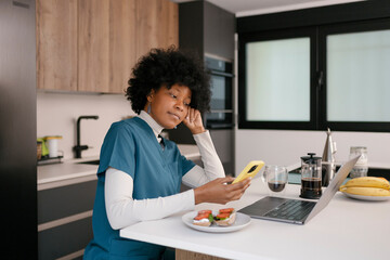 Healthcare worker having breakfast while checking smartphone and laptop