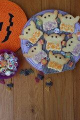 Top view of Halloweeen shortcrust cookies in shape of a ghost with white chocolate glaze and  sprinkles on a lilac plate with candies and pumpkin shape decoration on wooden table 