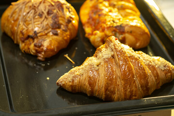 A high-angle close-up of three assorted golden-brown pastries, including a croissant and savory cheese-topped items, on a black baking tray.