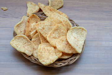 A high-angle close-up of golden, crispy Indonesian crackers (kerupuk) piled on a small wicker plate, set on a light wood surface.