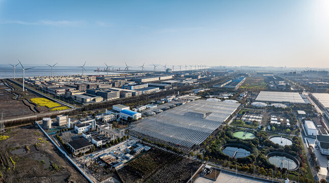 Aerial view of a coastal industrial zone featuring wind turbines for renewable energy and various factories under a clear sky.