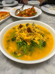 A high-angle view of Tumis Pocai Telur, an Indonesian/Chinese dish of pocai (spinach/amaranth) and multiple eggs (scrambled, salted, century) in a rich golden broth.