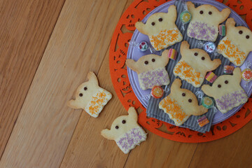 Top view of Halloweeen shortcrust cookies in shape of a ghost with white chocolate glaze and  sprinkles on a lilac plate on orange festive underplate  on wooden table 