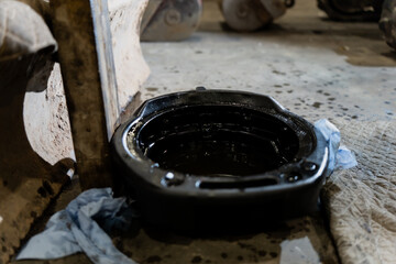 Oil drain catch pan positioned beneath heavy machinery with dark lubricants, service rags and workshop floor debris indicating maintenance procedures and equipment fluid collection tasks