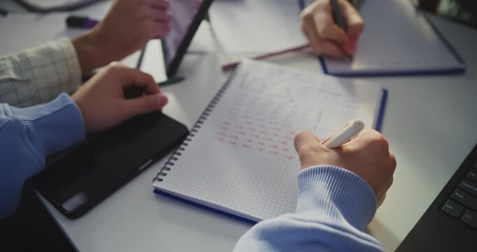 Close Up Hands of Several Students Intently Working at Shared Table. Student Writing in Notebook Next to Laptop Screen Displaying Analytical Graphs. Concept of Data Analysis, Digital Learning.