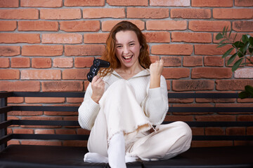 A young beautiful girl sits on the sofa holding a gamepad in her hands and playing a computer game. The player rejoices in victory