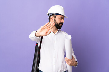 Young architect man with helmet and holding blueprints over isolated purple background making stop gesture and disappointed