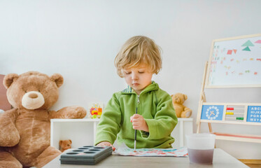 Cute little child boy sitting at table, painting on paper with paintbrush and colorful paints, funny and educational indoor activity for kids.