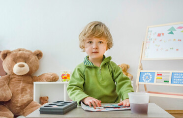Cute little child boy sitting at table, painting on paper with paintbrush and colorful paints, funny and educational indoor activity for kids.