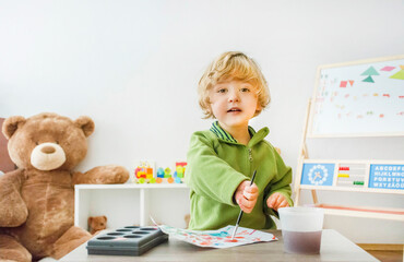 Cute little child boy sitting at table, painting on paper with paintbrush and colorful paints, funny and educational indoor activity for kids.