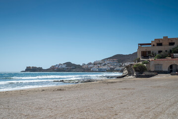 A serene beach in San Jose, Almeria, Spain, with golden sand, gentle waves, and white hillside houses. The blue sky and rocky coastline create a picturesque Mediterranean landscape