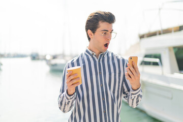 Young handsome man using mobile phone and holding a coffee with surprised expression