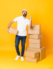 Full-length shot of delivery man among boxes over isolated yellow background surprised and pointing...