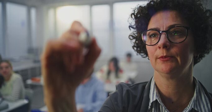 Close Up Adult Woman Wearing Glasses Actively Writes With Marker on Invisible Interactive Board. Students Listen Attentively. Concept of Modern Teaching Methods, Innovations in Educational Technology.
