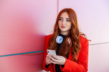 Naklejka premium A young woman wearing a red jacket and headphones stands against a colorful wall, looking at her phone. She appears focused and engaged with her device in a lively environment.