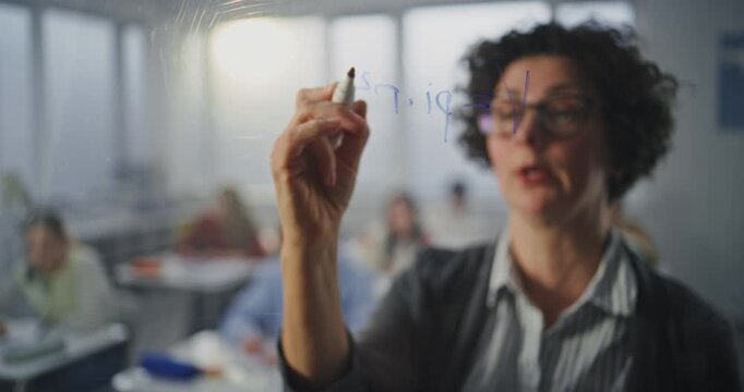 Dynamic Female Teacher With Glasses Writes With Marker Formulas, Addressing Class With Engaging Expression. Concept of Clear Communication, and Teacher Role in Fostering Student Comprehension.