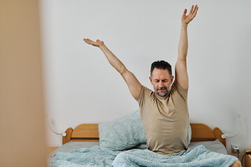 Middle-aged man relaxing and sleeping and waking up and stretching in bed on a cozy morning in bedroom