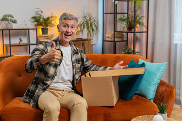 Happy young man unpacking delivery parcel sitting at home apartment on sofa. Smiling satisfied guy shopper online shop customer opening cardboard box receiving purchase gift by fast postal shipping.