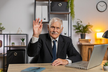 Hello. Happy senior businessman grandfather waves hand hi gesture greeting welcomes someone, online webinar, remote distance conversation. Freelancer in suit sitting at desk with laptop in home office