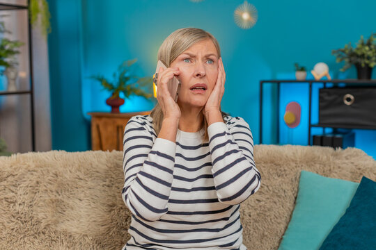 Mature upset anxious frustrated woman listening bad news talking on mobile phone. Grandmother sits on the sofa at home, holding the smartphone having conversation and showing signs of distress, stress - Powered by Adobe
