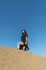 Young woman with dark hair wearing sunglasses poses confidently on sandy hill against clear blue sky, showcasing a sense of adventure and freedom in outdoor lifestyle