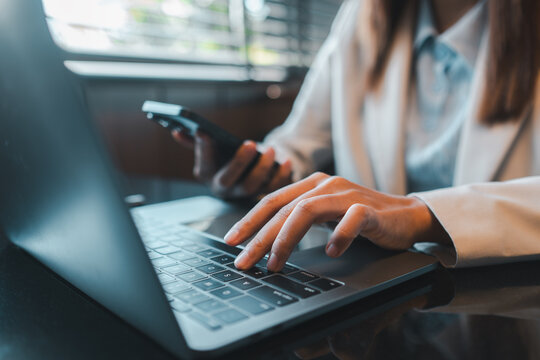 Close-up of a businesswoman multitasking with a smartphone and laptop in a modern office environment.