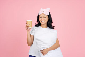 Asian woman in pajamas and an eye mask hugging a pillow with a cup of hot coffee on a pink background.