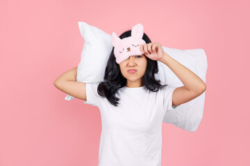 Asian woman in pajamas removing her blindfold and hugging a pillow behind her neck on a pink background.