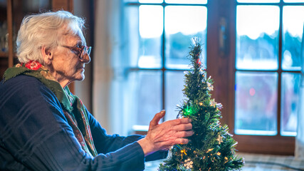Elderly woman decorating a Christmas tree at home with colorful ornaments and warm lights