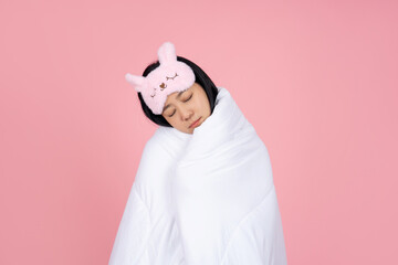 Smiling young woman in pajamas and sleep mask holding a white blanket, closing her eyes and resting at home. Relaxing lifestyle concept, good mood.