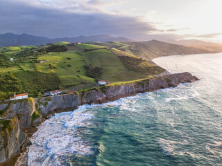 Aerial view of the Atlantic coastline with steep cliffs, green hills, and waves breaking at sunset, Basque Country, Spain