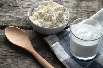 Fresh cottage cheese in a white bowl and a glass of kefir on a rustic wooden table, with a wooden spoon and checkered cloth creating a cozy, natural setting