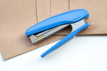 An open blue and silver handheld stapler rests on a stack of textured brown kraft paper envelopes or documents, set against a clean white surface, highlighting essential office supplies and organizati