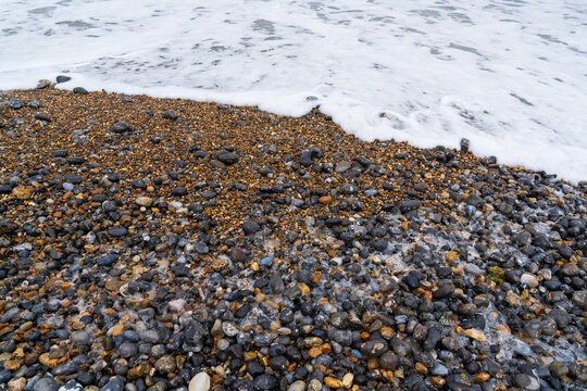 Colourful pebble beach with pounding ocean waves and white foam breaking on rocky shore. Natural coastal landscape displaying smooth stones in various hues and a dynamic seaside environment - Powered by Adobe
