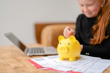Child saving coins in a yellow piggy bank with laptop nearby