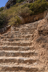 A rugged stone staircase carved into the rocky hillside at Preveli Beach, Crete. The ancient, weathered steps lead upward toward dense Mediterranean shrubs under bright summer sunlight. A natural