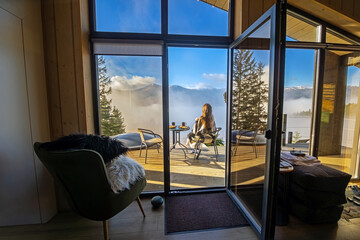Woman enjoying mountain view from cozy cabin. A woman wrapped in a blanket sits on a terrace, drinking coffee and admiring a breathtaking Mountain View above the clouds.