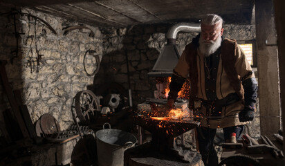 A Viking blacksmith with a white beard and braided hair works intently at a forge, hammering glowing metal on an anvil, with flames flickering in the background of a stone workshop.