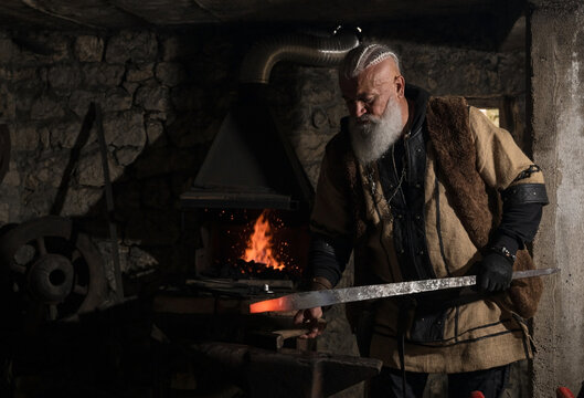 A Viking blacksmith with a white beard and braided hair works intently at a forge, hammering glowing metal on an anvil, with flames flickering in the background of a stone workshop.