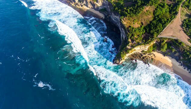 Aerial view turquoise ocean waves crashing on sandy beach, lush green cliffs above, bright sunny daylight - Powered by Adobe