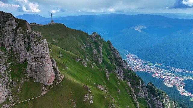 Distancing from the Caraiman Peak with a Cross memorial. Aerial view on the dangerous path on the green mountain. Romania, the Bucegi Mountains of the Southern Carpathians.