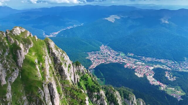Approaching the spectacular mountain peak with dangerous steep slopes. Aerial perspective on the residential area scattered in the valley surrounded by lush greenery. The Bucegi Mountains, Romania.