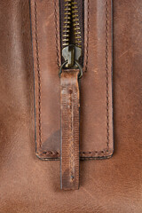 A close-up shot of a rustic brown leather texture with a brass zipper and a leather pull tab, showing worn details and stitching