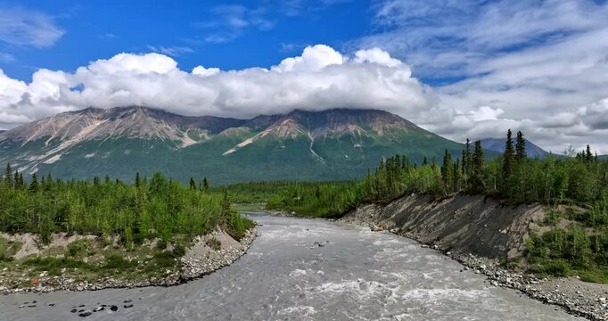 Rushing river below Alaska mountain range. Fast gray water carves a forested canyon at the foot of cloud wrapped Alaska peaks.