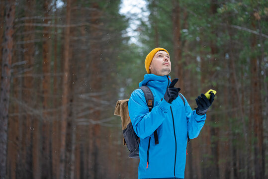 Man wearing winter clothing and backpack, looking up and pointing while holding a smartphone, actively seeking navigation or a signal outdoors in a cold, snowy, dense pine forest environment