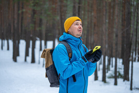 Man hiker wearing a blue jacket and yellow beanie feeling lost in a snow-covered forest, looking up and searching for direction while holding a smartphone and realizing disorientation