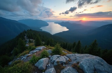 Scenic mountaintop view over lake valley at sunset. Sun sets on horizon above distant peaks, forested hills. Calm water reflects cloudy sky. Rocky ridge with evergreen trees in foreground. Serene